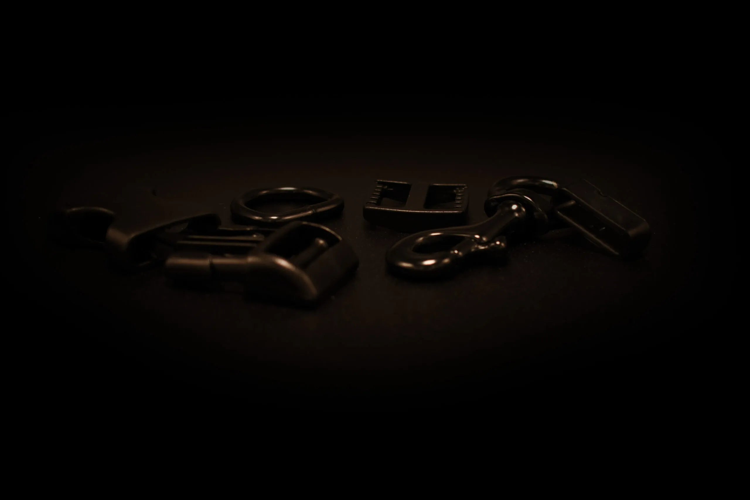 Verschiedene schwarze Metall- und Kunststoffbeschläge wie Schnallen, Karabiner und Verbindungselemente auf einer dunklen Oberfläche angeordnet