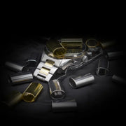 Verschiedene zylindrische Metallhülsen in Silber- und Goldoptik, die um eine Armbanduhr auf einer dunklen Oberfläche angeordnet sind