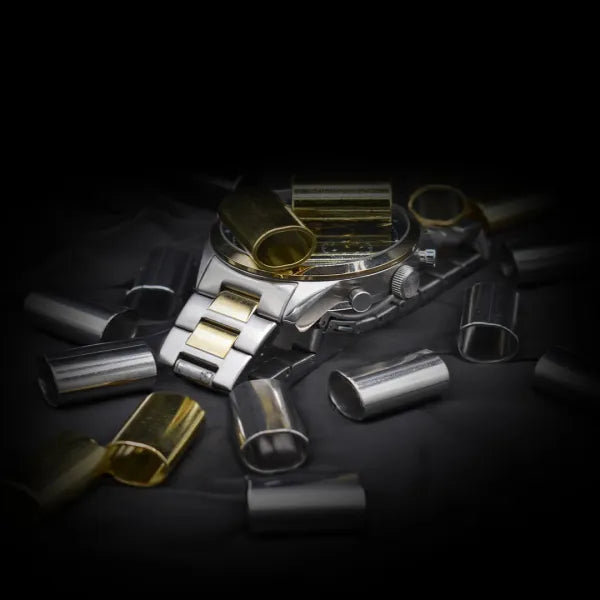 Verschiedene zylindrische Metallhülsen in Silber- und Goldoptik, die um eine Armbanduhr auf einer dunklen Oberfläche angeordnet sind